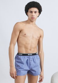 Lyseblå boxershorts laget av mykt stoff med en svart midjebånd som har «BOSS»-logoen. Front med knappelukking, avslappet passform.