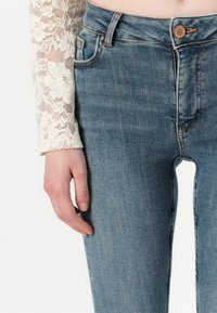 Vaqueros de denim azul claro con cintura alta, con costuras detalladas y un botón de cobre. Una mano reposa sobre el muslo, con una manga de encaje visible.