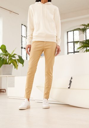 Person iført hvid hættetrøje, beige bukser og hvide sneakers, der står indendørs nær planter og et hvidt surfbræt med "Light"-logo.