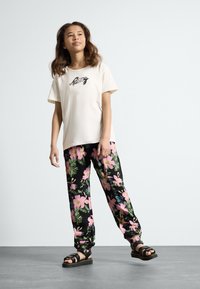Ljusbeige bomullströja med broderad text, i kombination med svarta byxor med en färgglad blommig mönstring, bärs med svarta sandaler.