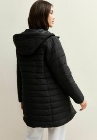 Giacca parka nera con cappuccio, caratterizzata da una texture trapuntata orizzontale, un taglio leggermente più lungo e polsini regolabili.