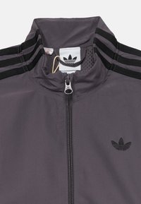 Siva zip-up jakna, izdelana iz lahkega materiala. Ima črne detajle, tri vzporedne črte na ramenih in logotip na prsih.