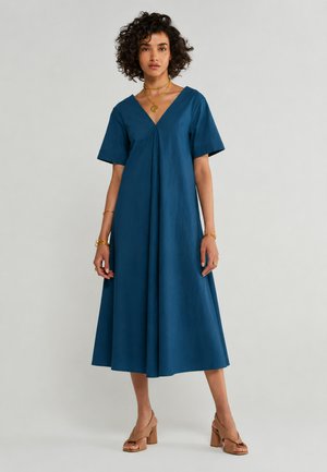 Donna con capelli ricci che indossa un abito midi blu con scollo a V, gioielli dorati e sandali marroni con tacco largo, in piedi davanti a uno sfondo uniforme.