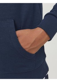 Hoodie bleu marine avec poignets et ourlet côtelés. Doté d'une poche kangourou à l'avant. Texture de tissu lisse. Gros plan sur une main dans la poche.