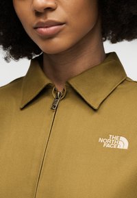 Κοντινό πλάνο ενός ατόμου που φοράει μια ελιά πράσινη ζακέτα με γιακά και φερμουάρ, με το λευκό λογότυπο The North Face στο στήθος.