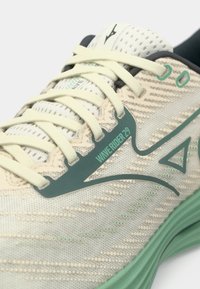 Baskets de course beige clair avec un supérieur en maille texturée, des accents verts et des lacets crème. Présente la marque "WAVE RIDER 29" sur le côté.