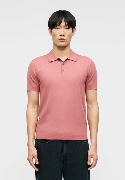 Jeune homme aux cheveux noirs raides portant un polo rose à manches courtes et un pantalon foncé, debout devant un fond clair uni.