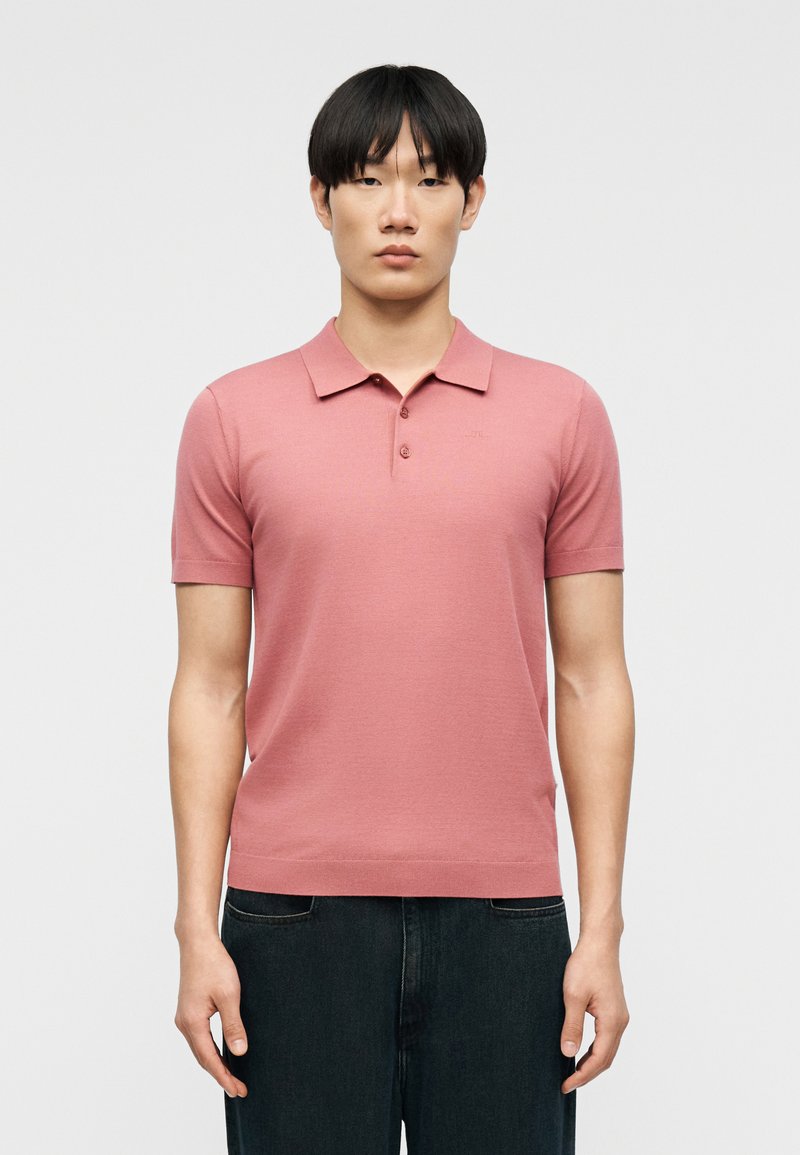 Νέος άνδρας με ίσια μαύρα μαλλιά, φορώντας κοντομάνικο ροζ πόλο και σκουρόχρωμο παντελόνι, στέκεται μπροστά σε ένα απλό φωτεινό φόντο.