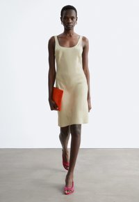 Femme marchant dans une robe beige sans manches tenant une pochette orange, portant des sandales à talons roses et des boucles d'oreilles créoles dorées.