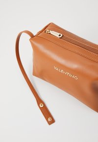 Lær kosmetikkpung i tan-farge med gull glidelås, med håndleddstropp og preget "VALENTINO"-logo på forsiden. Glatt tekstur.