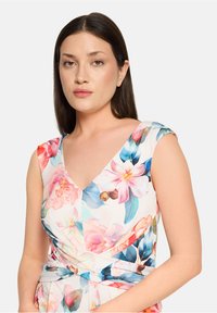 Femme aux cheveux raides et foncés, portant une robe blanche sans manches avec des motifs floraux roses et bleus, sur un fond clair uni.