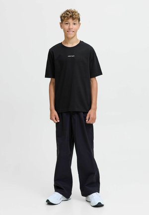 Zwarte t-shirt met korte mouwen en een wit logo op de borst, gecombineerd met losse zwarte broek en witte sneakers.