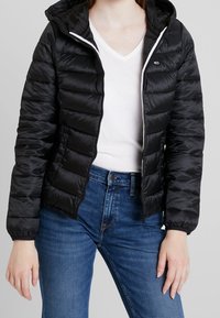 Czarna kurtkapuchowa z kapturem, z pikowanym wzorem, przednim zamkiem błyskawicznym i białym wykończeniem. Połączona z białą koszulą z dekoltem w serek i niebieskimi dżinsami.