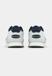 Baskets de sport blanches avec des accents bleu marine, présentant un talon rembourré et un branding visible "1973" à l'arrière. Upper texturé et semelle en caoutchouc.