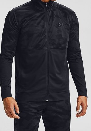 Homme portant une veste à fermeture éclair et un pantalon assorti à motif camouflage noir, debout devant un fond uni clair.