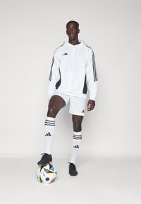 Weißer Kapuzenpullover mit schwarzen Akzenten, weiße Shorts und schwarze Fußballschuhe. Schwarz-weiße gestreifte Socken. Hält einen bunten Fußball.