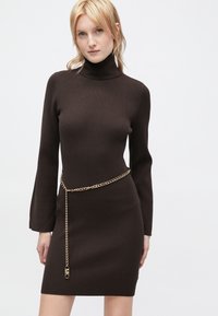Brun ribbet strikkjole med høj hals, lange ærmer og et guld kædebælte, der fremhæver taljen. Glat tekstur, tætsiddende design.