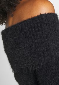 Pull noir à épaules dénudées avec texture côtelée, en matériau doux et duveteux. Le design met l'accent sur les épaules avec une coupe décontractée.