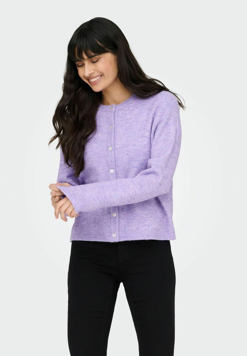 Cardigan lavande en tissu tricoté doux et texturé, doté d'une patte de boutonnage sur le devant et de manches longues, assorti à un pantalon noir.