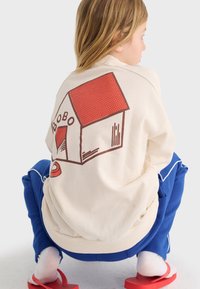 Niño agachado, vistiendo una sudadera blanca con un gráfico de caseta de perro en rojo y marrón, y pantalones azules con rayas blancas.