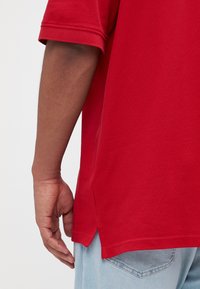 Rød bomulds t-shirt med korte ærmer, en buet kant med sidesplit, og en afslappet pasform, parret med lyseblå jeans.