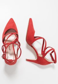 Escarpins rouges en daim avec bouts pointus, dotés de brides croisées et d'un talon fin. Semelle intérieure métallique lisse et design minimaliste.