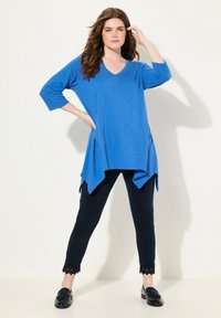 Blaues, kurzärmliges V-Ausschnitt-Tunika mit einem längeren hinteren Saum, kombiniert mit schwarzen Leggings mit Spitze an den Bündchen.