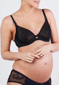 Fekete kismama melltartó átlátszó kosarakkal és hímzett virágmintákkal. Állítható pántokkal és kapcsos záródással rendelkezik, hozzáillő bugyival párosítva.