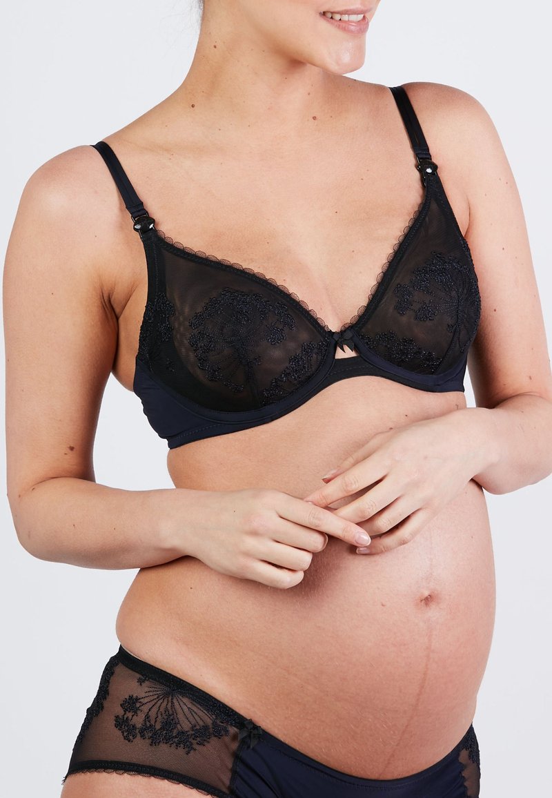 Fekete kismama melltartó átlátszó kosarakkal és hímzett virágmintákkal. Állítható pántokkal és kapcsos záródással rendelkezik, hozzáillő bugyival párosítva.