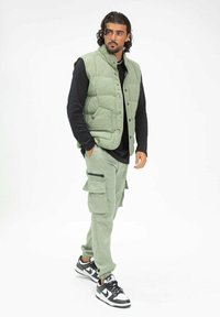 Veste matelassée vert clair sur un t-shirt noir à manches longues, associée à un pantalon cargo vert clair et des baskets noir et blanc, se tenant sur un fond blanc.