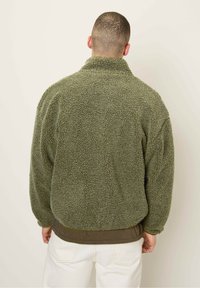 Chaqueta de forro polar verde con un cuello alto, superficie texturizada y un ajuste holgado. Llevada sobre una capa marrón y combinada con pantalones blancos.