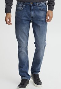 Vaqueros de mezclilla azul oscuro con un corte regular, diseño de cinco bolsillos, ligero desgaste y detalles cosidos. Llevados con zapatillas negras.