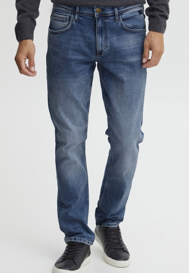 Vaqueros de mezclilla azul oscuro con un corte regular, diseño de cinco bolsillos, ligero desgaste y detalles cosidos. Llevados con zapatillas negras.