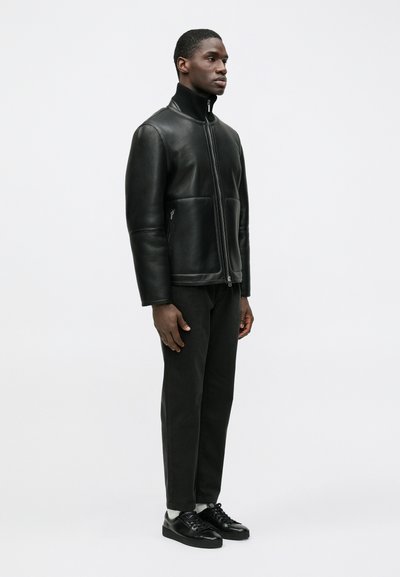 Schwarze Lederjacke mit hohem Kragen, enganliegendem Schnitt und seitlichen Reißverschlusstaschen, kombiniert mit schwarzer Hose und Schuhen, vor einfarbigem Hintergrund.