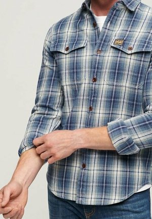 Homme retroussant la manche d'une chemise à carreaux bleue et blanche avec deux poches poitrine et des boutons marron.