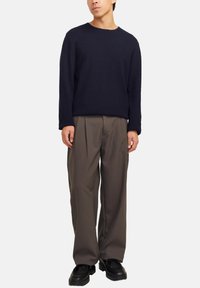 Männliches Modell trägt einen dunkelblauen Langarmpullover, lockere braune Faltenhose und schwarze Schuhe und steht vor einem weißen Hintergrund.