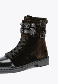 Black velvet ankle boot with a glossy black toe, decorative crystal accents, dual buckle straps, and a textured sole.
