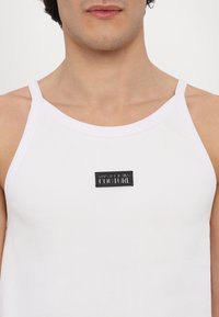 Hvit ribbet tanktop med brede stropper og en rektangulær svart lapp som står "VERSACE JEANS COUTURE." Glatt tekstur, tettsittende design.