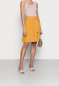 Kvinna som bär en ärmlös blus i spets med rosa kant, en senapsgul kjol med knytbälte, vita klacksandaler och håller en liten rosa handväska.