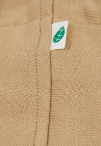 Beige kötött anyag, látható varrással, kicsi fehér címkével, zöld levél mintával a széle közelében. Puha tapintás, laza megjelenés.