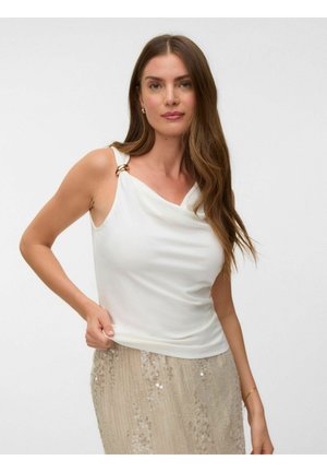 Femme aux longs cheveux bruns porte un haut sans manches blanc avec un détail anneau doré et une jupe beige à sequins sur fond blanc.
