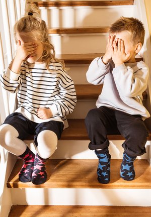 Two children sitting on wooden stairs covering their eyes, wearing casual clothes and star-patterned socks.