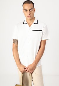 Short-sleeve white shirt with black accents on the collar and pocket, made of a smooth fabric. Model holds a beige bag.