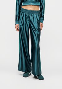 Μοντέλο που φοράει τιρκουάζ σατέν παντελόνια φαρδιάς γραμμής με ελαστική μέση και ταιριαστά τιρκουάζ αθλητικά παπούτσια, με cropped μπουφάν ορατό στο ύψος της μέσης.