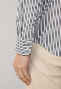 Camisa de vestir a rayas con líneas verticales azules y blancas, con puños abotonados y una textura de tela suave, combinada con pantalones beige claros.