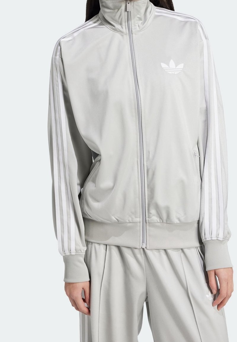 Žena oblečená v světle šedém adidas teplákovém soupravě s bílými pruhy na rukávech a kalhotách, se zipovou bundou a žebrovanými manžetami.