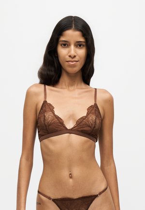 Femme aux longs cheveux foncés portant un bralette en dentelle marron et une lingerie en dentelle assortie, debout devant un fond blanc uni.
