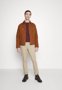 Braune Baumwolljacke mit Kontrastnähten, darüber ein kastanienbraunes Sweatshirt, beige Hosen und braune Stiefel, steht vor einem schlichten Hintergrund.