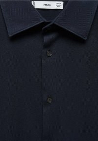 Chemise texturée bleu marine à manches courtes avec un col, présentant une patte de boutonnage avec deux boutons visibles, étiquetée "MNG".