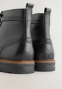 Čierne kožené členkové čižmy s brogue detailami, hrubými čiernymi gumovými podrážkami a hnedým prešívaním strednej podrážky, zobrazené zozadu.